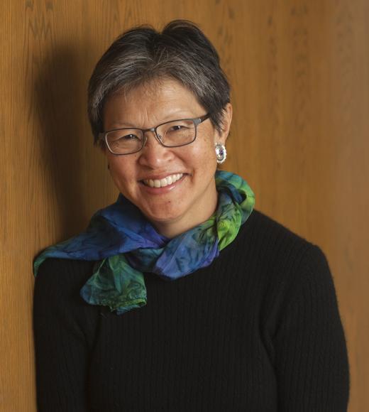 Smiling woman with short gray hair, glasses and blue-green scarf against wood backdrop