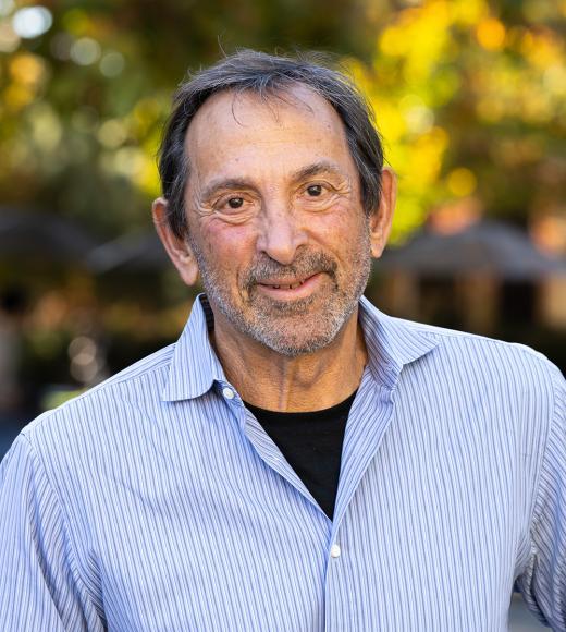 Portrait photograph of a smiling older man in a striped shirt with a sunlit leafy background