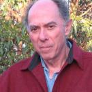 Balding older man in red jacket outdoors, neutral expression, leafy background