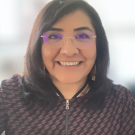 Smiling woman with dark hair, glasses and patterned zip jacket, blurred background photograph