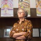 Older woman with glasses and patterned blouse stands arms crossed beneath colorful art in hallway.