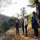 A group of five people stands on a dirt trail in a sunlit forested canyon, listening to one person speak while the others observe and engage. Sunlight filters through trees, and the terrain is dry and hilly with patches of green vegetation.